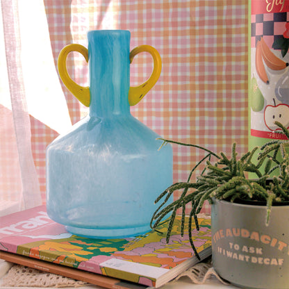 A blue vase with yellow double handles. The vase has a wide cylindrical base that tapers into a slender neck at the top. It is placed on a table in front of a pink striped background wall. To the right of the vase is a green plant.