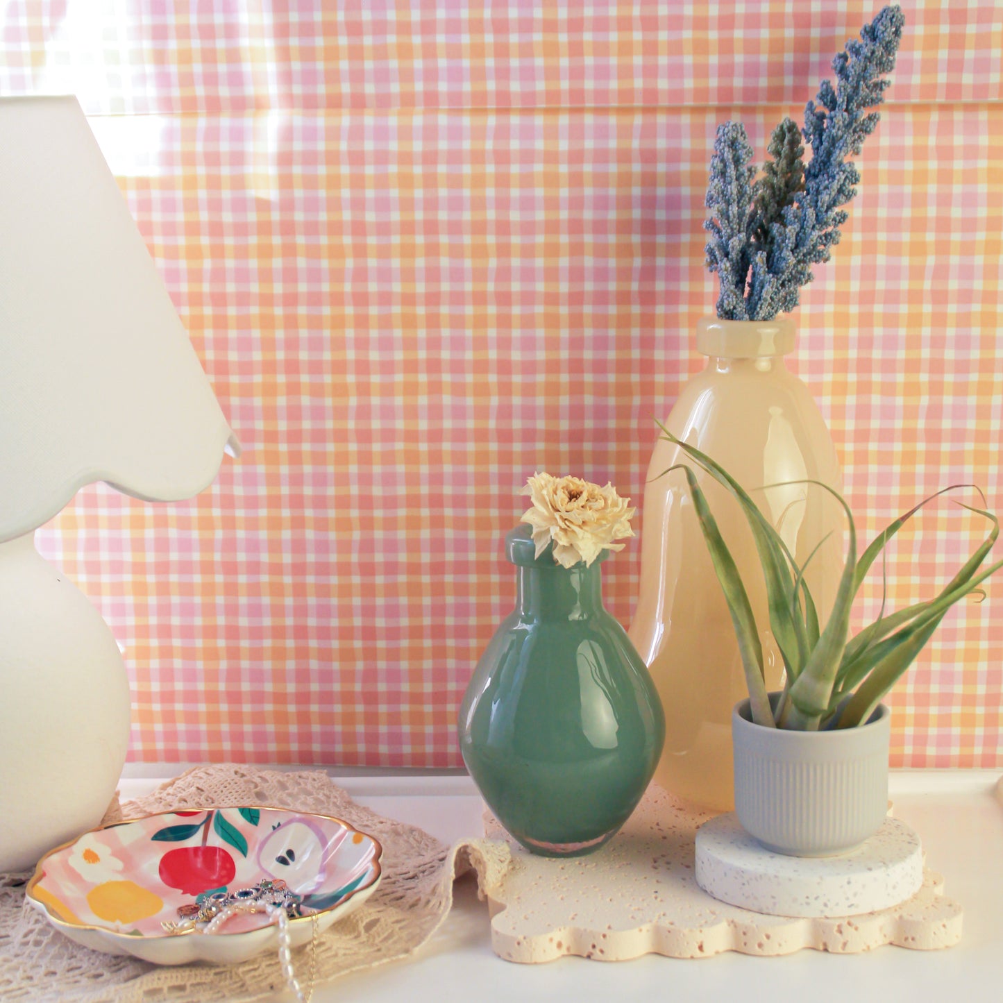 On a table set against a pink checkered background wall, a white table lamp is placed on the far left, with a jewelry tray in the center. On the far right, a light yellow gourd-shaped glass vase holds a bundle of purple lavender. In front of the vase, to its left, is a dark green round-bellied vase, and to its right sits an air plant.