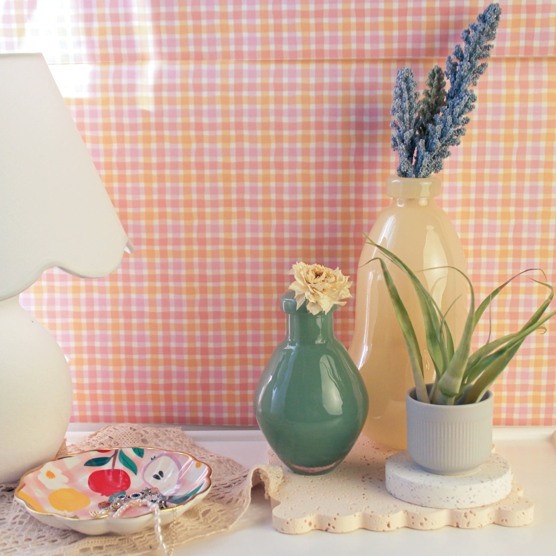 On a table set against a pink checkered background wall, a white table lamp is placed on the far left, with a jewelry tray in the center. On the far right, a light yellow gourd-shaped glass vase holds a bundle of purple lavender. In front of the vase, to its left, is a dark green round-bellied vase, and to its right sits an air plant.