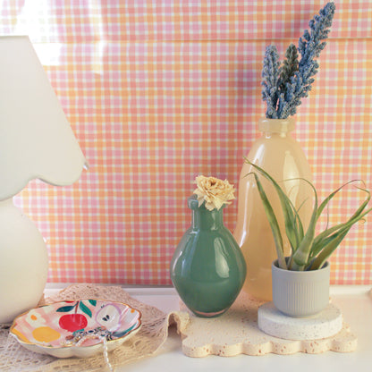 On a table set against a pink checkered background wall, a white table lamp is placed on the far left, with a jewelry tray in the center. On the far right, a light yellow gourd-shaped glass vase holds a bundle of purple lavender. In front of the vase, to its left, is a dark green round-bellied vase, and to its right sits an air plant.