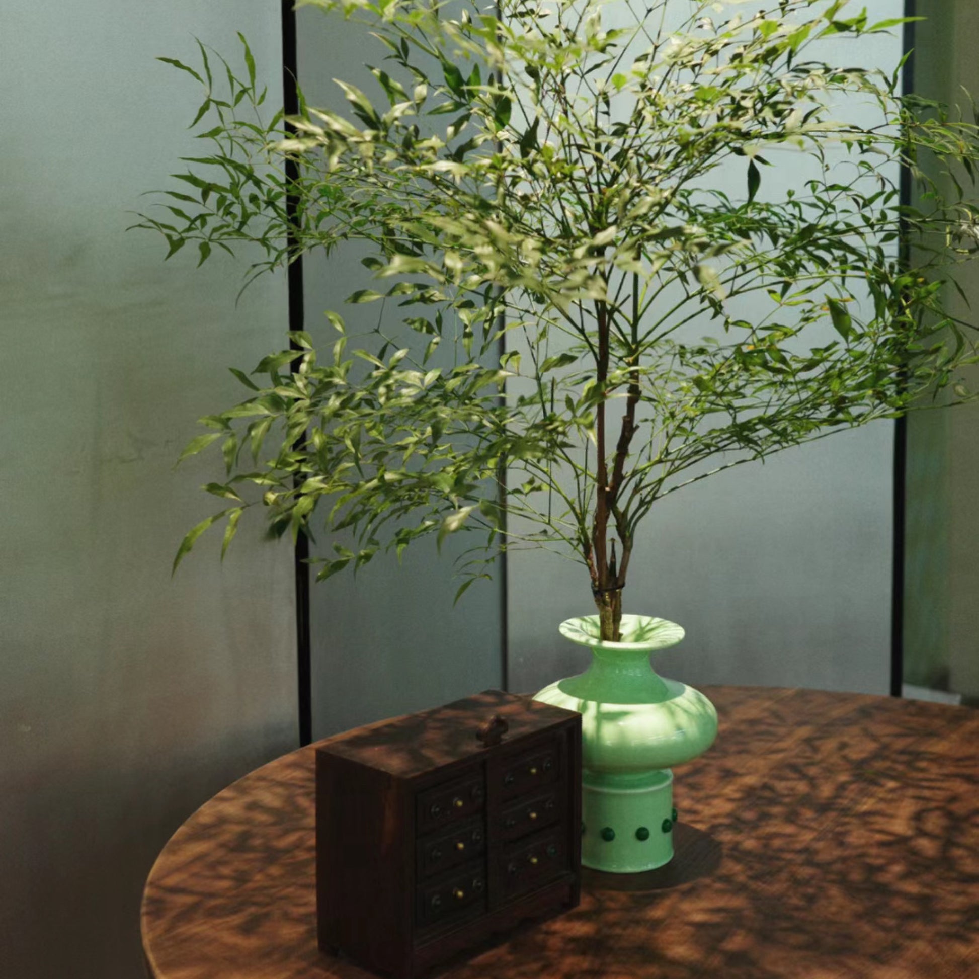 A brown wooden round table in front of a screen features a Chinese classical bamboo-shaped green vase inlaid with green jade, holding a vibrant bouquet of green branches. Next to the vase, a small wooden storage box is placed, adding a touch of rustic charm to the setting.