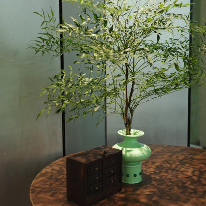A brown wooden round table in front of a screen features a Chinese classical bamboo-shaped green vase inlaid with green jade, holding a vibrant bouquet of green branches. Next to the vase, a small wooden storage box is placed, adding a touch of rustic charm to the setting.