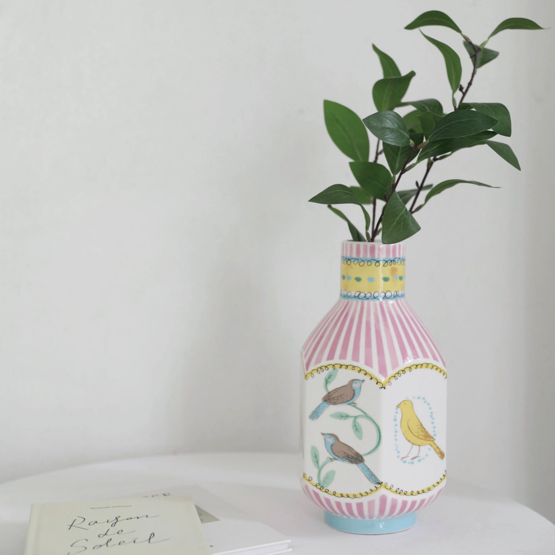 An octagonal narrow-neck vase rests on a tabletop, with its body hand-painted with nightingales in various colors and pink vertical stripes adorning the surface. Inside the vase, a green branch is elegantly arranged, while a book is placed to the left of the vase, completing the thoughtful and artistic display.