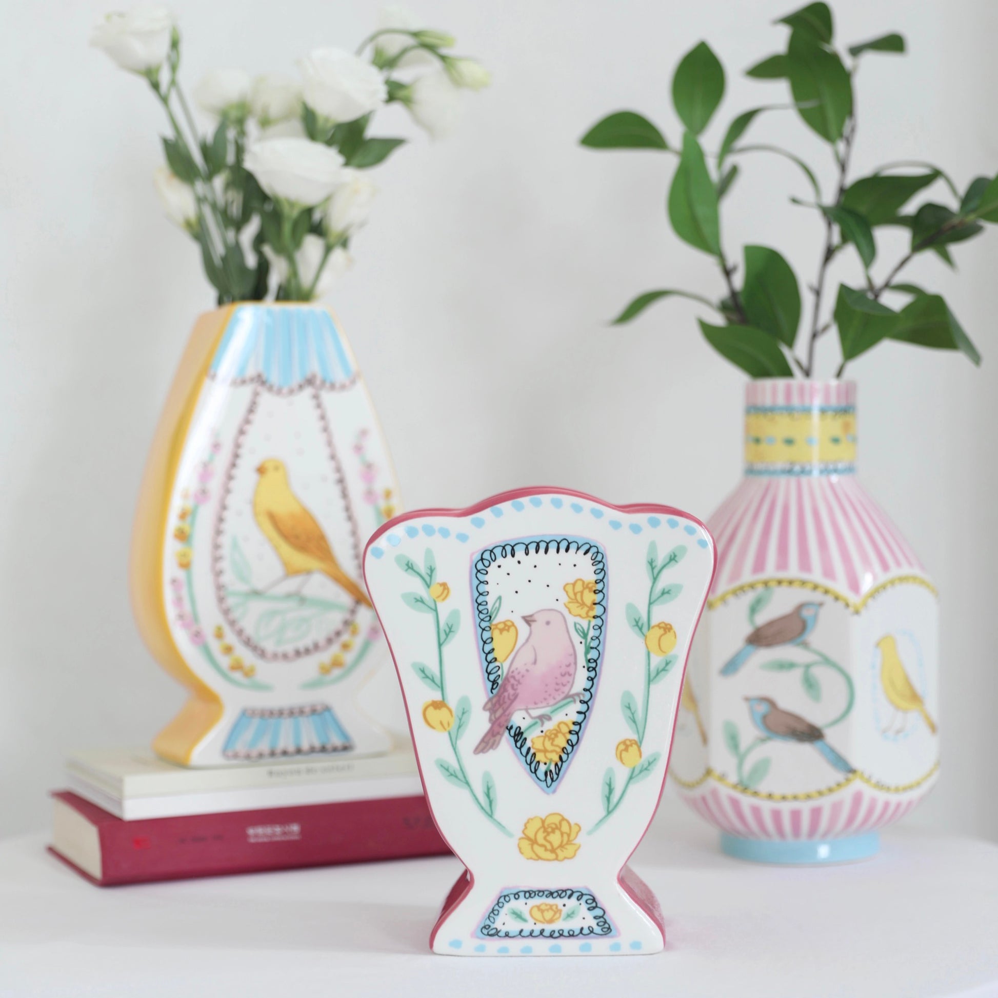 Three vases of different shapes are arranged in a staggered horizontal line. On the left, a rose bud-shaped vase with a bud is placed on a book, featuring a hand-painted yellow nightingale in the center and a yellow-colored side. Inside, a bouquet of white carnations is arranged. The middle vase is tulip-shaped, adorned with pink nightingales and yellow roses on its surface. On the right, a narrow-necked, octagonal vase is hand-painted with nightingales in various colors, with pink vertical lines