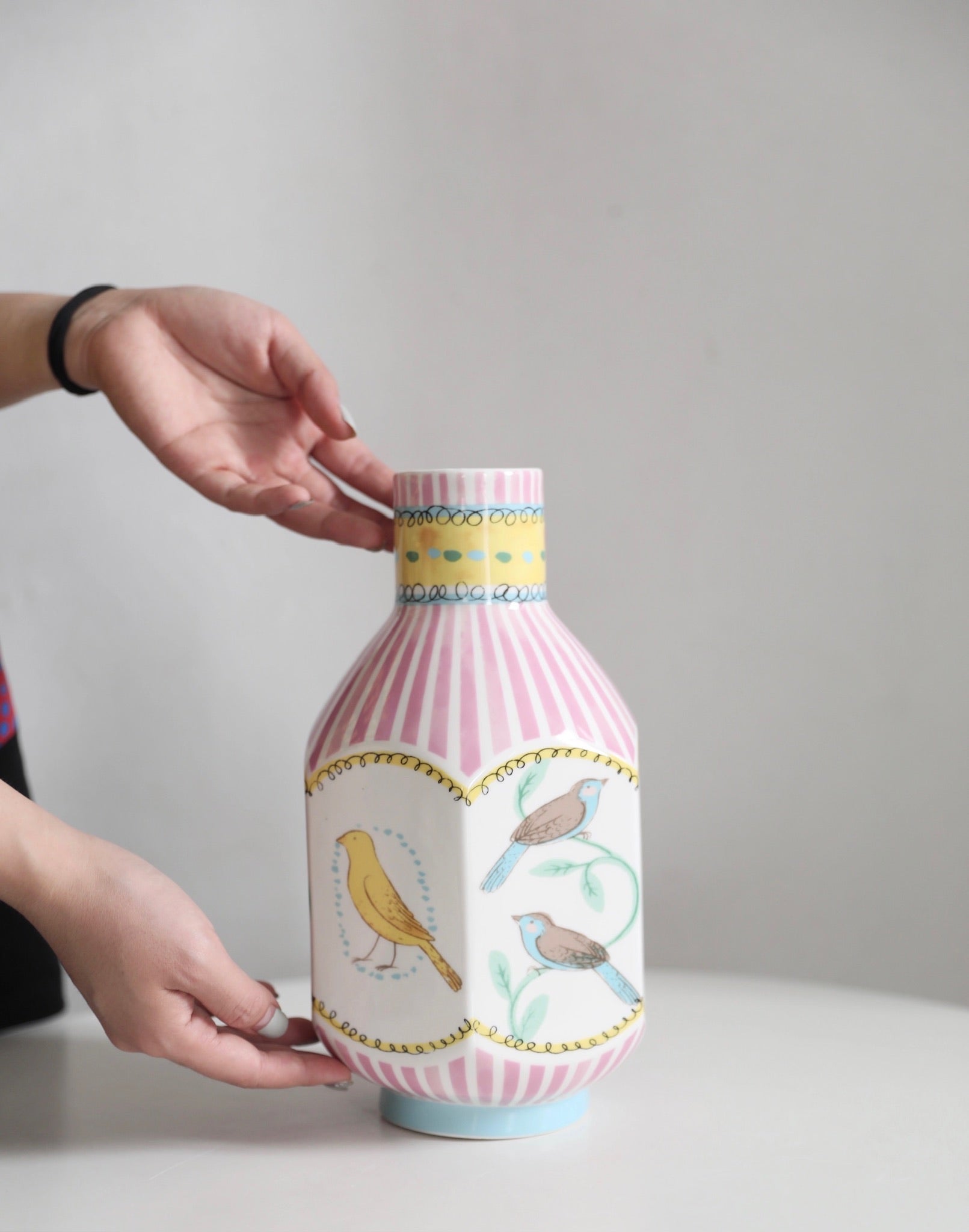 A pair of hands gently holding an octagonal narrow-neck vase on a table. The vase features hand-painted nightingales in various colors, with pink vertical stripes elegantly covering the surface, adding a sophisticated and artistic touch to the display.