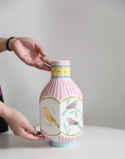 A pair of hands gently holding an octagonal narrow-neck vase on a table. The vase features hand-painted nightingales in various colors, with pink vertical stripes elegantly covering the surface, adding a sophisticated and artistic touch to the display.